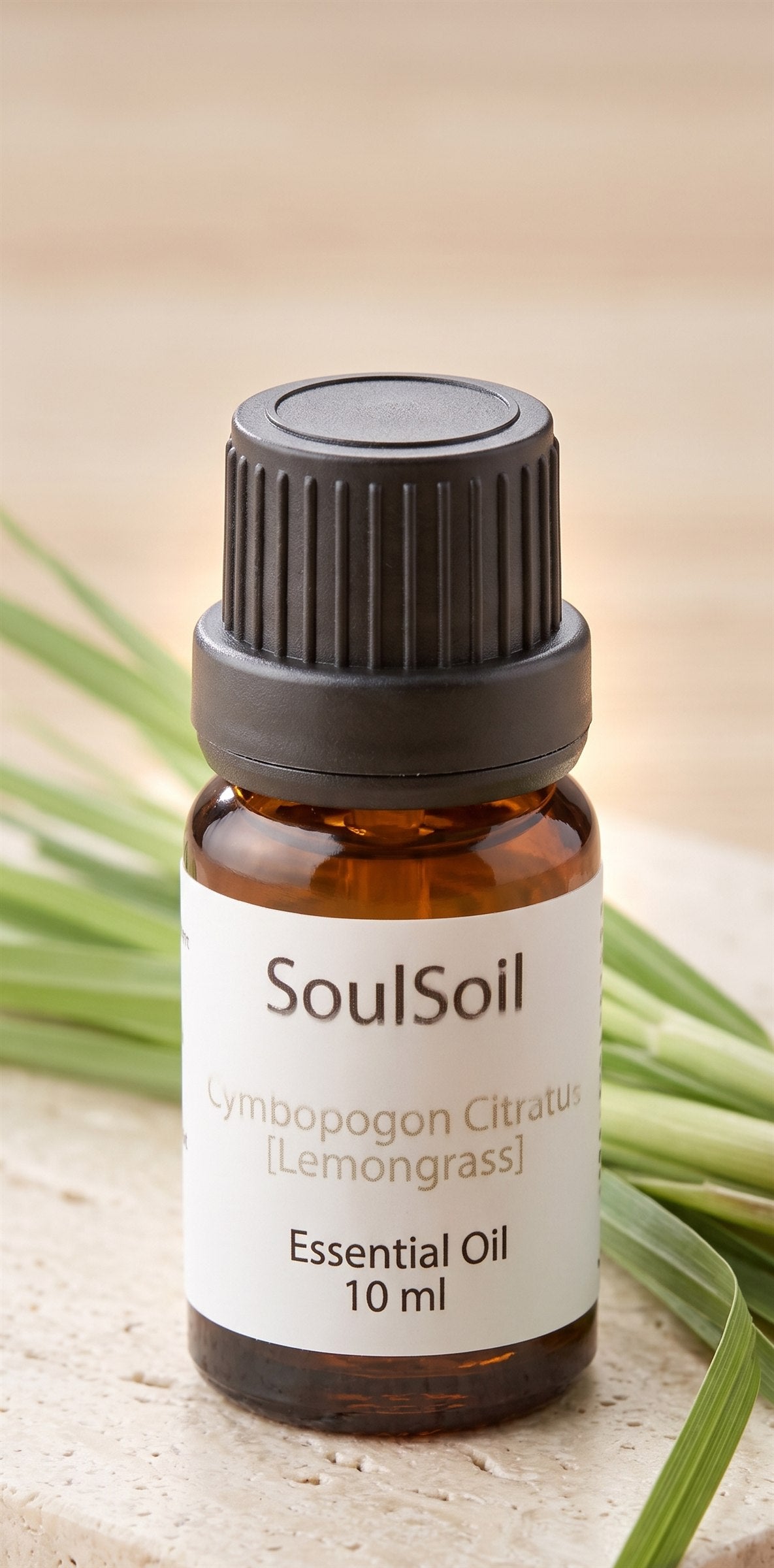 Close-up of a SoulSoil lemongrass bottle, labelled Cymbopogon citratus, 10 ml, on pale stone against fresh lemongrass blades.